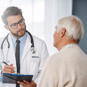 A doctor in a white coat with a stethoscope around his neck holds a clipboard and speaks attentively with an elderly male patient.